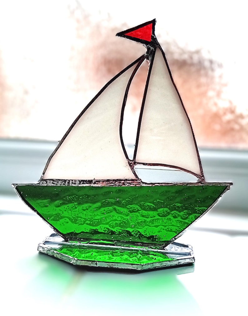 May include: A stained glass sailboat with a red flag, white sails, and a textured green hull. The boat sits on a clear, faceted base. The sailboat is a decorative nautical item.
