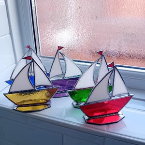 May include: A group of stained glass sailboat ornaments. Each boat has a white sail, a red flag, and a base in different colours: gold, purple, green, red, and blue. The sailboats are arranged on a white surface.