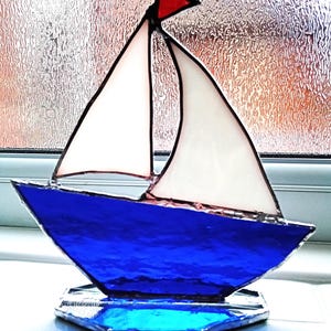 May include: A stained glass sailboat ornament featuring a deep blue hull, two white sails, and a small red triangular flag at the top. The pieces are joined by dark metal solder lines, and the entire structure rests on a blue glass base.