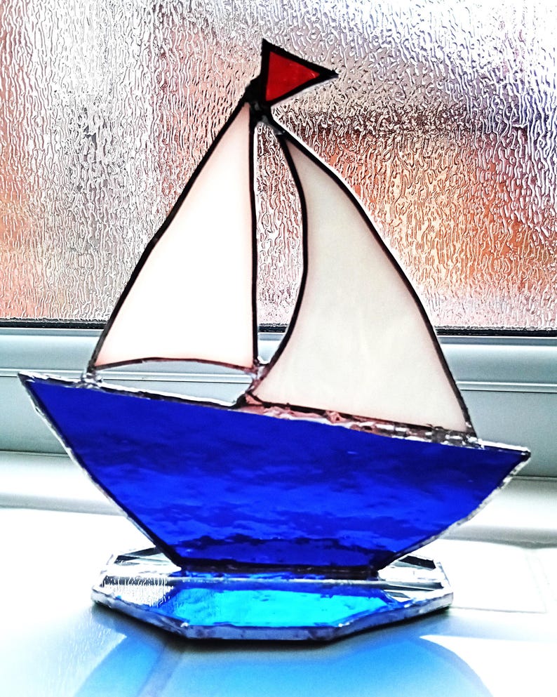 May include: A stained glass sailboat ornament featuring a deep blue hull, two white sails, and a small red triangular flag at the top. The pieces are joined by dark metal solder lines, and the entire structure rests on a blue glass base.