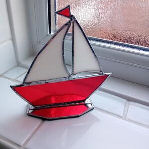 May include: A stained glass model of a sailboat. The boat features a red hull, white sails, and a small red flag. The glass is framed with a dark metal. The boat is displayed on a white surface.