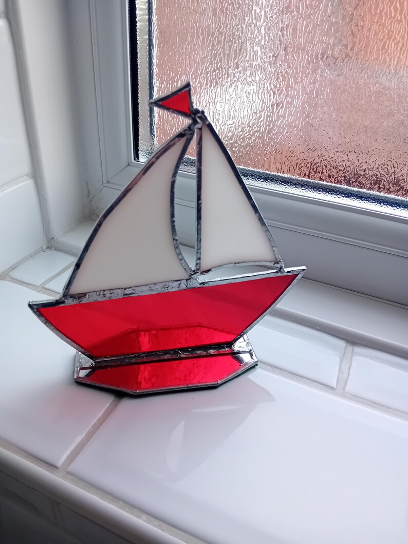 May include: A stained glass model of a sailboat. The boat features a red hull, white sails, and a small red flag. The glass is framed with a dark metal. The boat is displayed on a white surface.