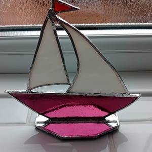 May include: A stained glass sailboat ornament featuring a vibrant pink hull and base with white sails and a small red flag at the top. The glass pieces are joined together with silver-coloured metal solder lines, creating a clear, defined structure.