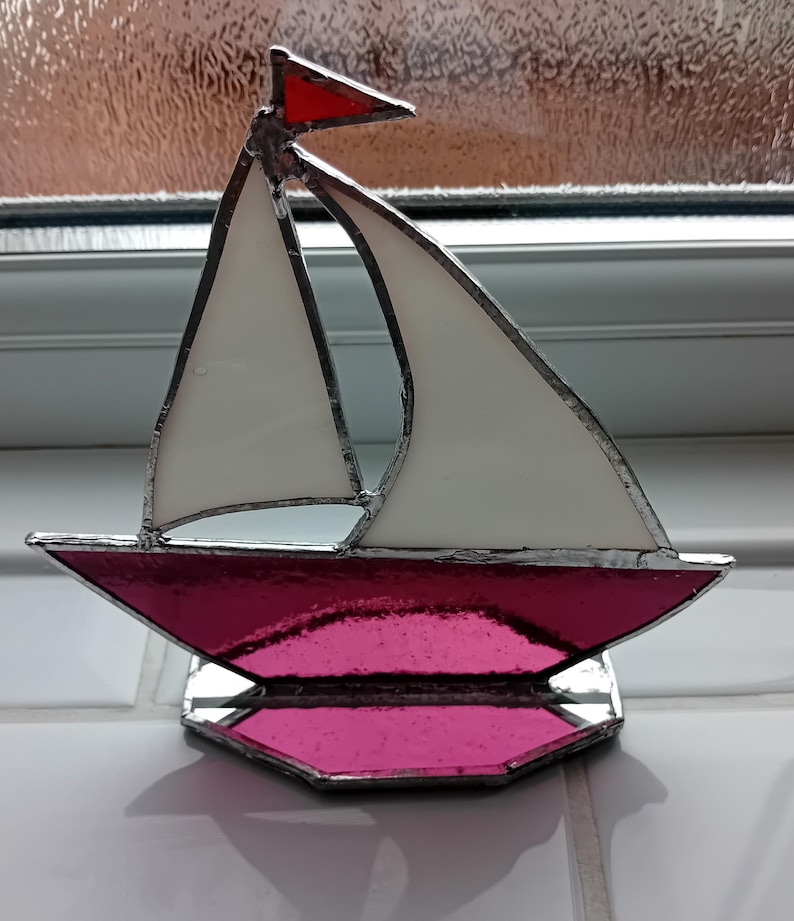 May include: A stained glass sailboat ornament featuring a vibrant pink hull and base with white sails and a small red flag at the top. The glass pieces are joined together with silver-coloured metal solder lines, creating a clear, defined structure.