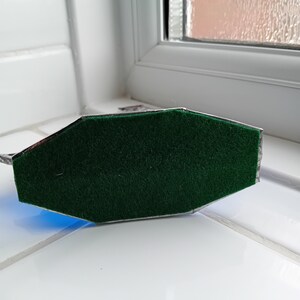 May include: An octagonal base covered in a vibrant green felt material. The base is framed by a thin, metallic silver border that outlines its geometric shape, providing a clean edge to the soft, textured green surface.