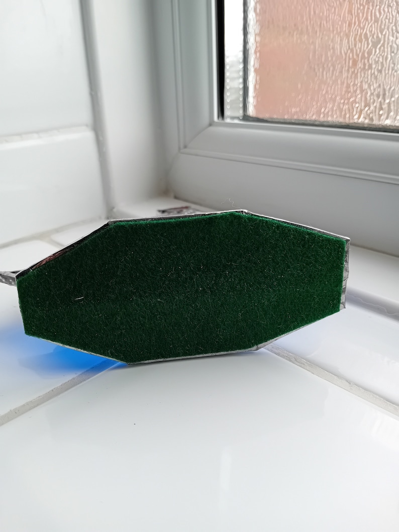 May include: An octagonal base covered in a vibrant green felt material. The base is framed by a thin, metallic silver border that outlines its geometric shape, providing a clean edge to the soft, textured green surface.