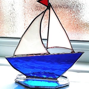 May include: A decorative stained glass sailboat. The boat features a bright blue hull and base, white sails, and a red pennant. The glass reflects light, creating a shimmering effect. The sailboat is a nautical decorative item.