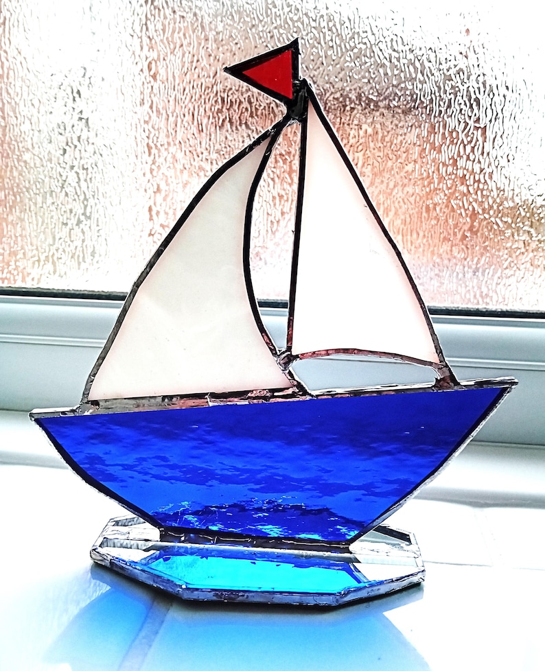 May include: A decorative stained glass sailboat. The boat features a bright blue hull and base, white sails, and a red pennant. The glass reflects light, creating a shimmering effect. The sailboat is a nautical decorative item.
