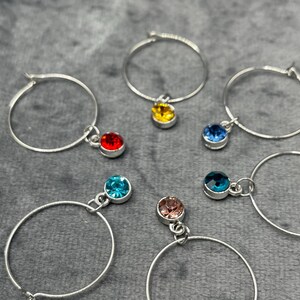 May include: Silver hoop earrings with colorful gemstone charms. The charms are in various colors, including red, yellow, blue, and pink. The earrings are displayed on a gray surface.