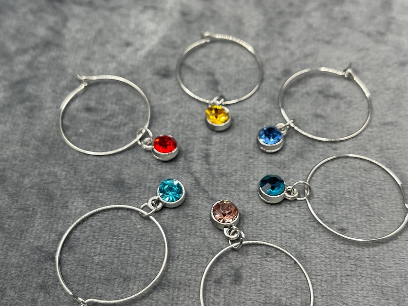 May include: Silver hoop earrings with colorful gemstone charms. The charms are in various colors, including red, yellow, blue, and pink. The earrings are displayed on a gray surface.