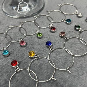 May include: A collection of silver wine glass charms with colorful gemstone pendants. The charms are circular and feature various jewel tones, including red, blue, green, pink, and yellow. A wine glass is visible in the background.
