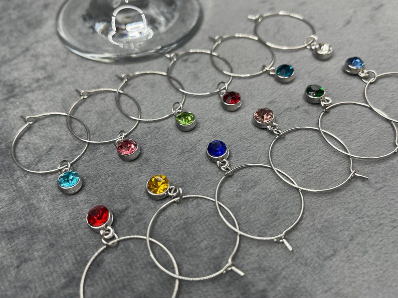May include: A collection of silver wine glass charms with colorful gemstone pendants. The charms are circular and feature various jewel tones, including red, blue, green, pink, and yellow. A wine glass is visible in the background.