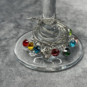 May include: A set of silver wine glass charms with colorful gemstone pendants. The charms are arranged on a wine glass, with a variety of jewel tones including red, yellow, blue, and green. The charms are made of metal.