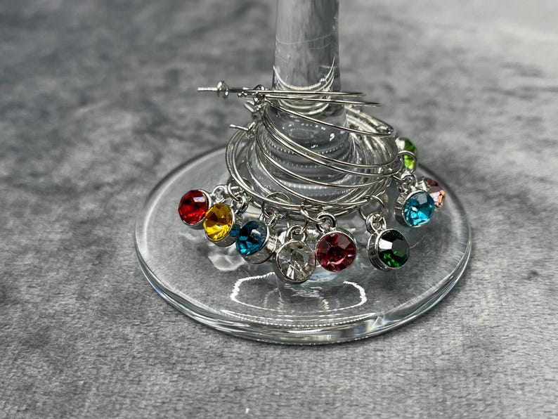 May include: A set of silver wine glass charms with colorful gemstone pendants. The charms are arranged on a wine glass, with a variety of jewel tones including red, yellow, blue, and green. The charms are made of metal.
