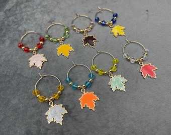 Puede incluir: Una colección de dijes para copas de vino con aros dorados, cada uno con un dije de hoja de arce de diferente color. Los dijes tienen varias cuentas de colores, incluyendo rojo, verde, azul y amarillo. Las hojas están pintadas en tonos de rosa, amarillo, marrón, naranja y verde.