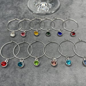 May include: A collection of silver wine glass charms with colorful gemstone pendants. The charms are arranged on a gray surface, with a wine glass in the background. Each charm features a different colored gem, including red, blue, green, and yellow.