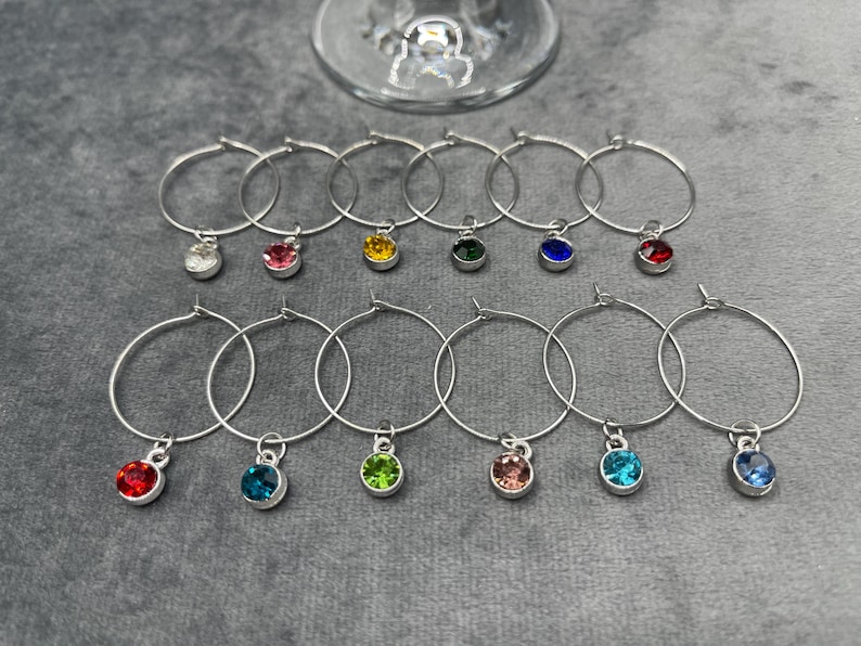 May include: A collection of silver wine glass charms with colorful gemstone pendants. The charms are arranged on a gray surface, with a wine glass in the background. Each charm features a different colored gem, including red, blue, green, and yellow.