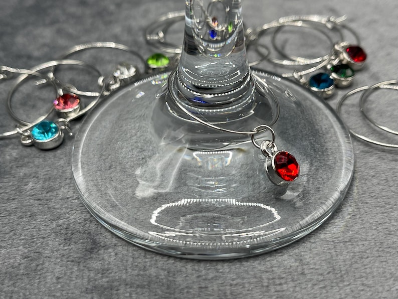 May include: A close-up of a wine glass with several silver wine charms. One charm is attached to the glass and features a red gemstone. Other charms have blue, green, and pink gemstones.
