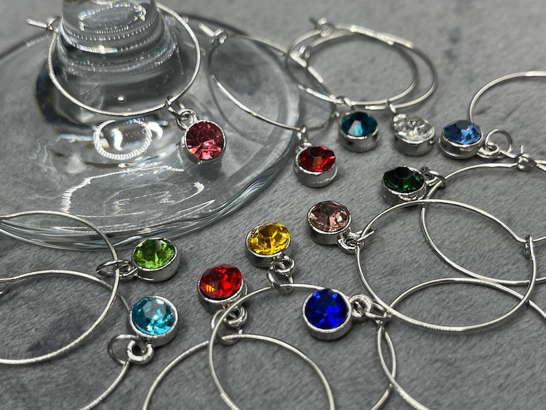 May include: A collection of silver wine glass charms with colorful gemstone pendants. The charms feature a variety of jewel tones, including red, blue, green, yellow, and pink. The charms are displayed near a wine glass.