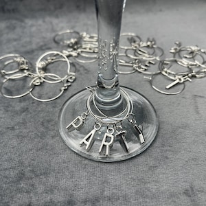 May include: Silver wine glass charms spelling out the word "PARTY". The charms are hanging from a wine glass.