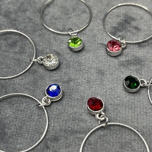 May include: Silver hoop earrings with colorful gemstone charms. The earrings feature a variety of colored gemstones, including clear, green, pink, blue, red, and dark green. The earrings are displayed on a gray surface.