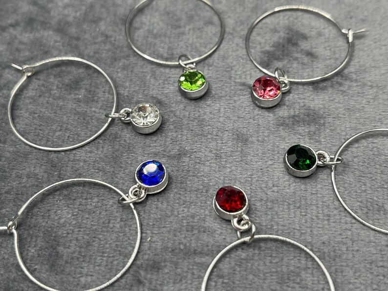 May include: Silver hoop earrings with colorful gemstone charms. The earrings feature a variety of colored gemstones, including clear, green, pink, blue, red, and dark green. The earrings are displayed on a gray surface.