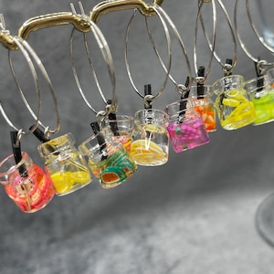 May include: A collection of silver hoop earrings featuring miniature clear glass jar charms. Each jar contains colourful fruit slice decorations and a small black straw. The earrings are displayed hanging from a gold-toned metal rack.