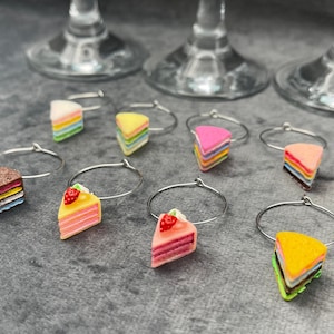 May include: A selection of cake slice wine charms. Each charm depicts a miniature cake slice in various colours and designs, attached to a silver-tone metal ring. The charms are displayed on a grey surface, with wine glasses in the background.