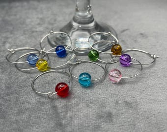 May include: Set of eight silver wine glass charms with colorful glass beads. The beads are red, yellow, blue, green, purple, pink, and two shades of blue.
