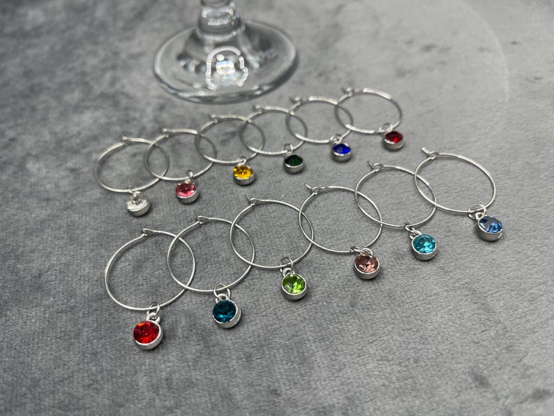 May include: A collection of silver wine glass charms with colorful gemstone pendants. The charms are arranged around the base of a wine glass. Each charm features a different colored gemstone, including red, blue, green, and pink.