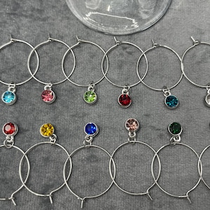 May include: A collection of silver-toned wine charms with colorful gemstone pendants. Each charm features a circular hoop with a small, round gem in various colors, including blue, pink, green, and red. The charms are arranged on a gray surface.