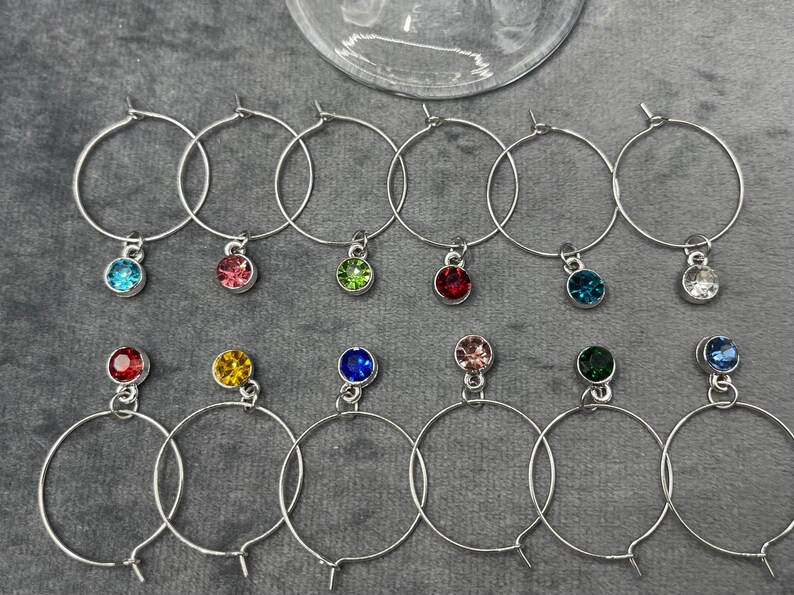 May include: A collection of silver-toned wine charms with colorful gemstone pendants. Each charm features a circular hoop with a small, round gem in various colors, including blue, pink, green, and red. The charms are arranged on a gray surface.