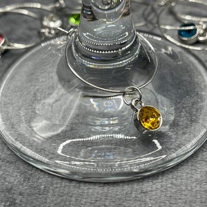 May include: Close-up of a wine glass with a silver charm and a yellow gemstone. Other wine charms with different colored gemstones are visible in the background. The glass is clear and sits on a gray surface.