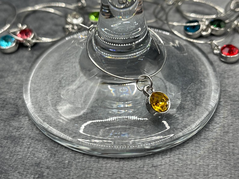 May include: Close-up of a wine glass with a silver charm and a yellow gemstone. Other wine charms with different colored gemstones are visible in the background. The glass is clear and sits on a gray surface.