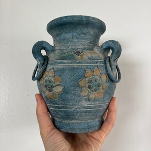 May include: A small, blue ceramic vase with two handles and a floral design. The vase has a distressed, vintage look.