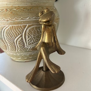 May include: A gold-coloured metal figurine of a frog sitting on a round base. The frog has a wide smile and is sitting with its legs crossed.