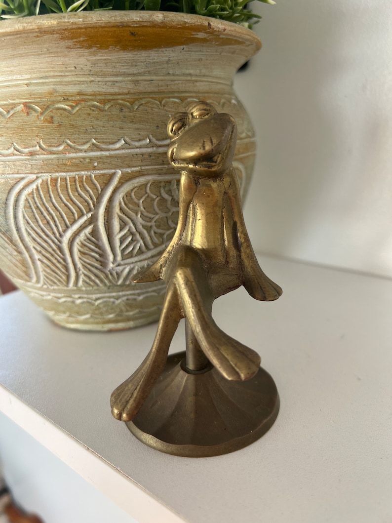 May include: A gold-coloured metal figurine of a frog sitting on a round base. The frog has a wide smile and is sitting with its legs crossed.