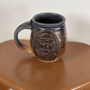 Peut inclure: Une tasse en céramique brune et bleue avec un motif amérindien. La tasse a une anse et un corps arrondi. Le motif représente une coiffe amérindienne avec des plumes et les mots "Eagle Rock Pottery".
