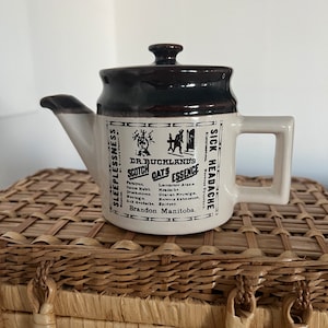 May include: A brown and white ceramic teapot with a lid. The teapot has a black and white label that reads "Dr. Buckland's Scotch Oats Essence" and lists various ailments it is said to cure. The label also includes the text "Brandon Manitoba".