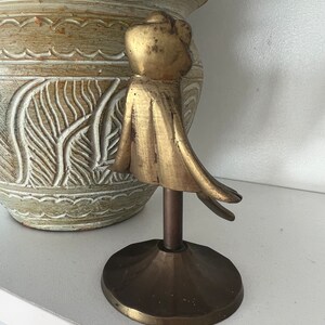 May include: A gold metal decorative stand with a round top and three legs. The stand features a stylised figure of a person with a flowing cape.