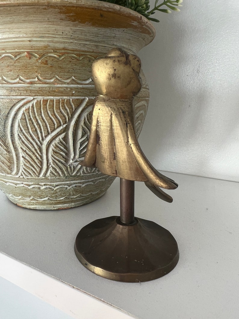 May include: A gold metal decorative stand with a round top and three legs. The stand features a stylised figure of a person with a flowing cape.