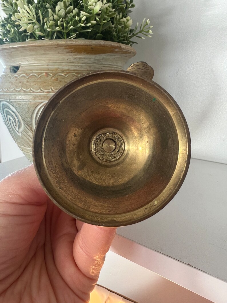 May include: Brass candle holder with a round base and a decorative design. The base is smooth and shiny.