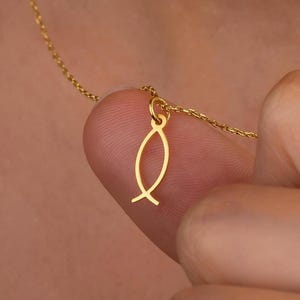 Puede incluir: Un collar dorado con un colgante de pez. El colgante es un contorno estilizado de un pez. La cadena del collar es delicada y fina. El colgante se sostiene entre dos dedos.