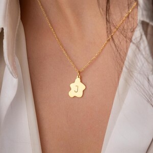 Peut inclure: Un collier doré avec un pendentif en forme de fleur. Le pendentif porte la lettre "J". Le collier est porté par une personne vêtue d'une chemise blanche.