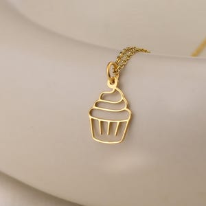 Puede incluir: Un collar dorado con un colgante de cupcake. El colgante es un contorno delicado de un cupcake con un diseño detallado de glaseado. El collar se muestra sobre una superficie de tonos neutros, resaltando el diseño de la joya.