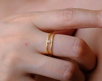 Puede incluir: Un anillo de oro con una pequeña piedra transparente engastada en el centro. El anillo se lleva en el dedo anular de una mano.