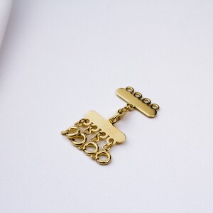 May include: Gold-toned metal jewelry clasp with multiple loops. The clasp has a flat, rectangular shape with a decorative edge and a row of five loops on one side. The other side has a row of five loops that hang down.