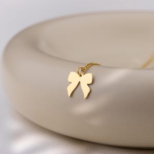 May include: A gold-colored bow pendant necklace. The pendant is a simple bow shape, and the chain is delicate. The necklace is displayed on a cream-colored, curved surface. The lighting is soft, highlighting the necklace's details.