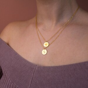 May include: A gold necklace with two pendants. The top pendant is a heart shape and the bottom pendant is a round disc with the letter "S" engraved on it.