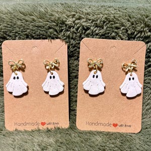 Pode incluir: Dois pares de brincos de fantasma brancos feitos à mão com olhos de pontos pretos e detalhes de laço dourado. Cada par é preso a um cartão castanho com o texto "Handmade with love". Os brincos são exibidos em uma superfície verde texturizada.